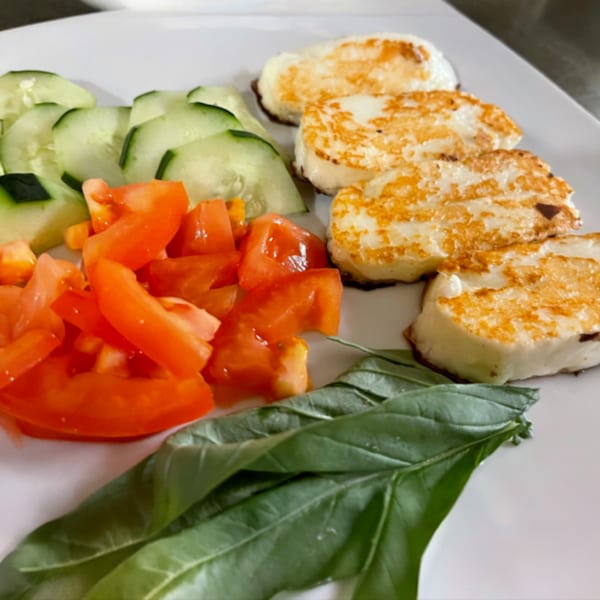 Grilled halloumi cheese with sliced cucumbers, diced tomatoes, and fresh basil leaves on a white plate.