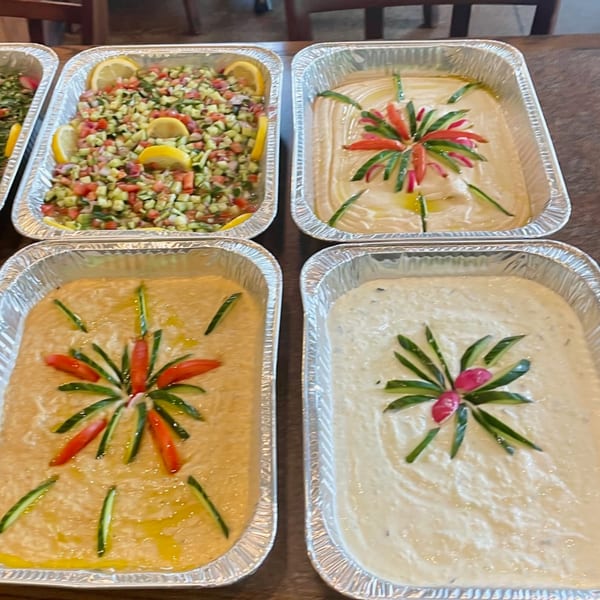 Four foil trays of Lebanese dips and salads, including hummus, baba ghanoush, and chopped salad.