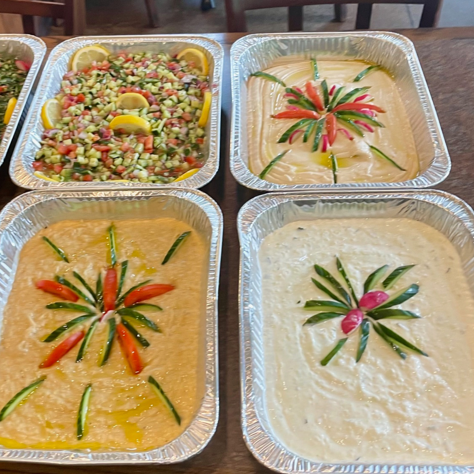 Four foil trays of Lebanese dips and salads, including hummus, baba ghanoush, and chopped salad.