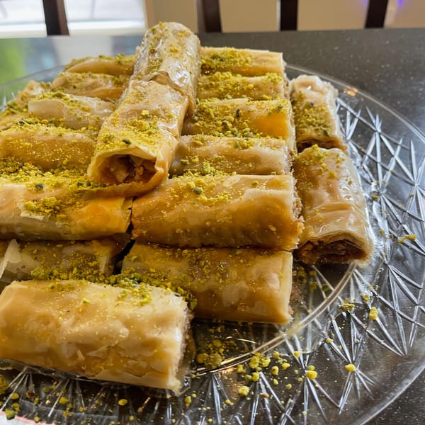 A clear glass plate filled with golden, syrup-drizzled Lebanese baklava rolls, generously sprinkled with green pistachios.