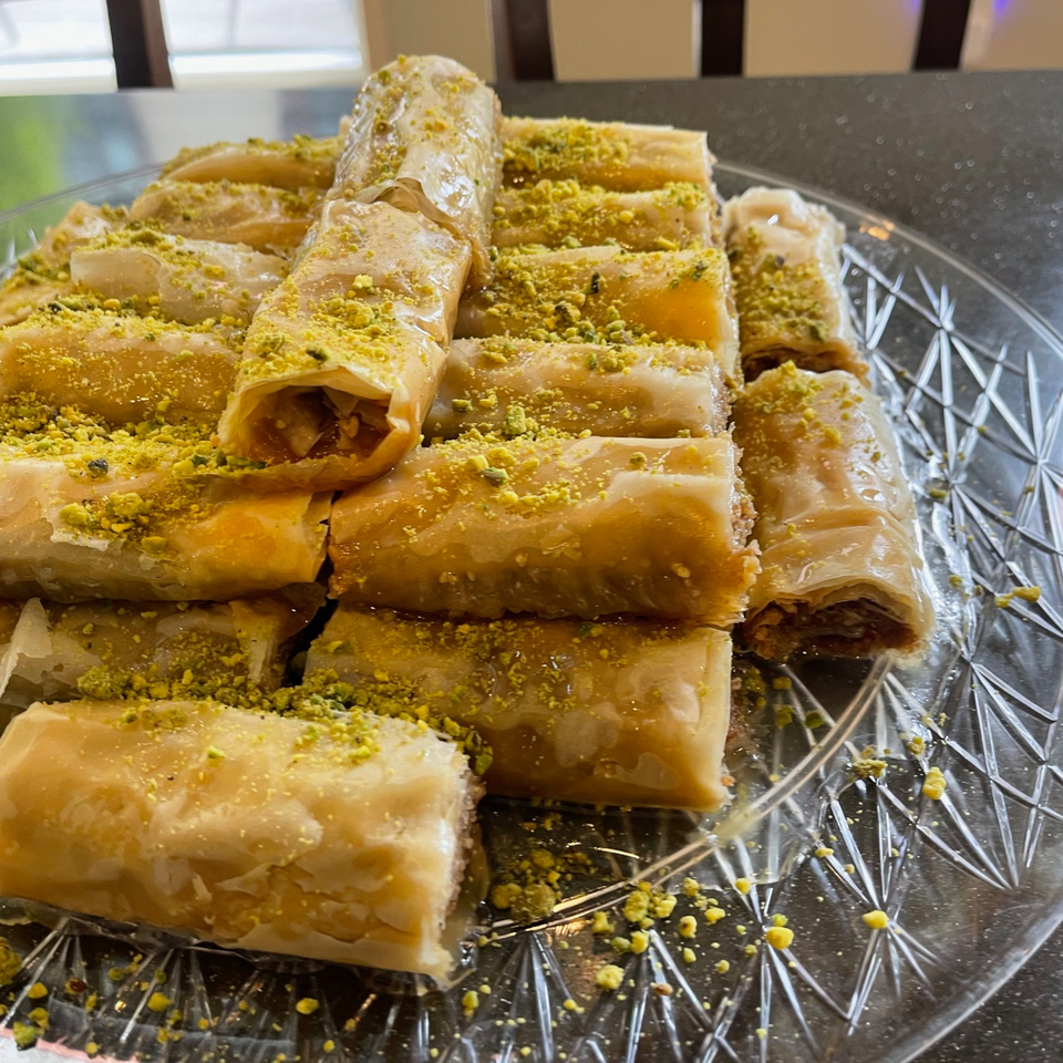 A clear glass plate filled with golden, syrup-drizzled Lebanese baklava rolls, generously sprinkled with green pistachios.